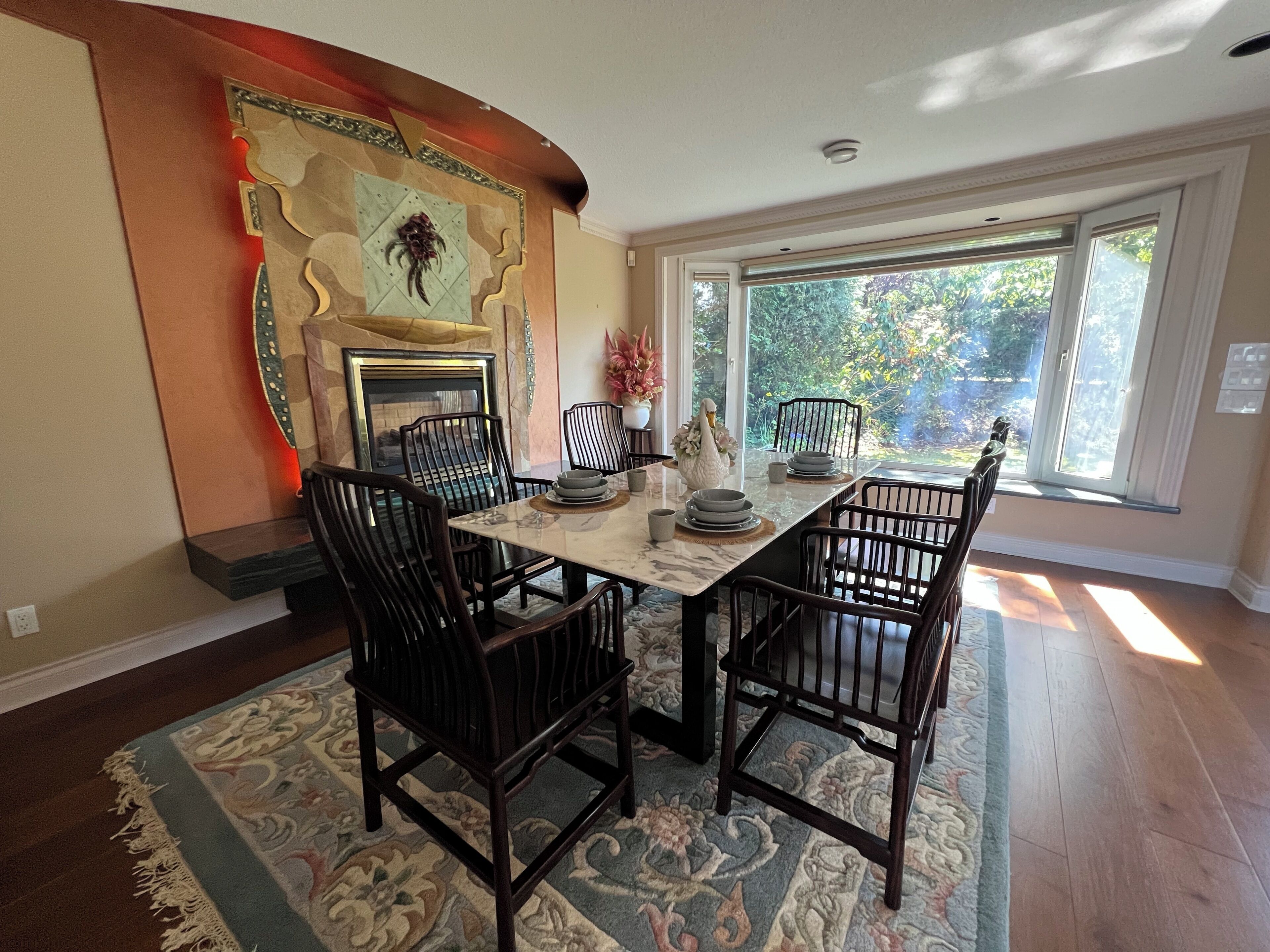 A dining area with a marble table, dark wooden chairs, a fireplace, and a view of greenery through large windows.