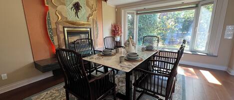 A dining area with a marble table, dark wooden chairs, a fireplace, and a view of greenery through large windows.
