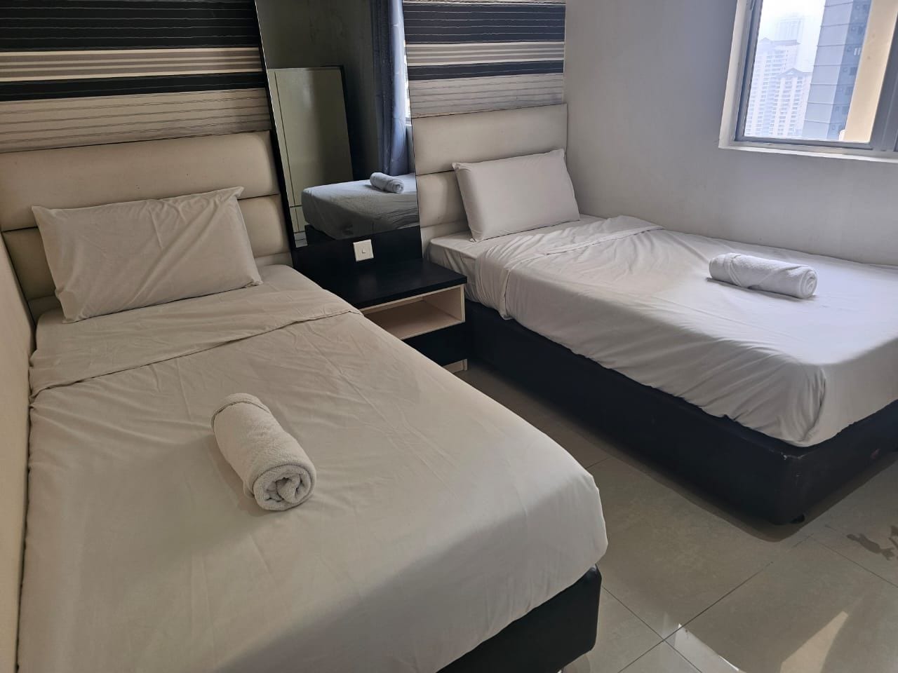 A hotel room with two beds, a nightstand, and a window with a view of buildings.