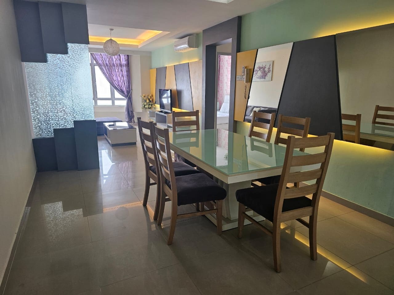 A modern dining area with a glass table and wooden chairs, a television, and a view into the living area.