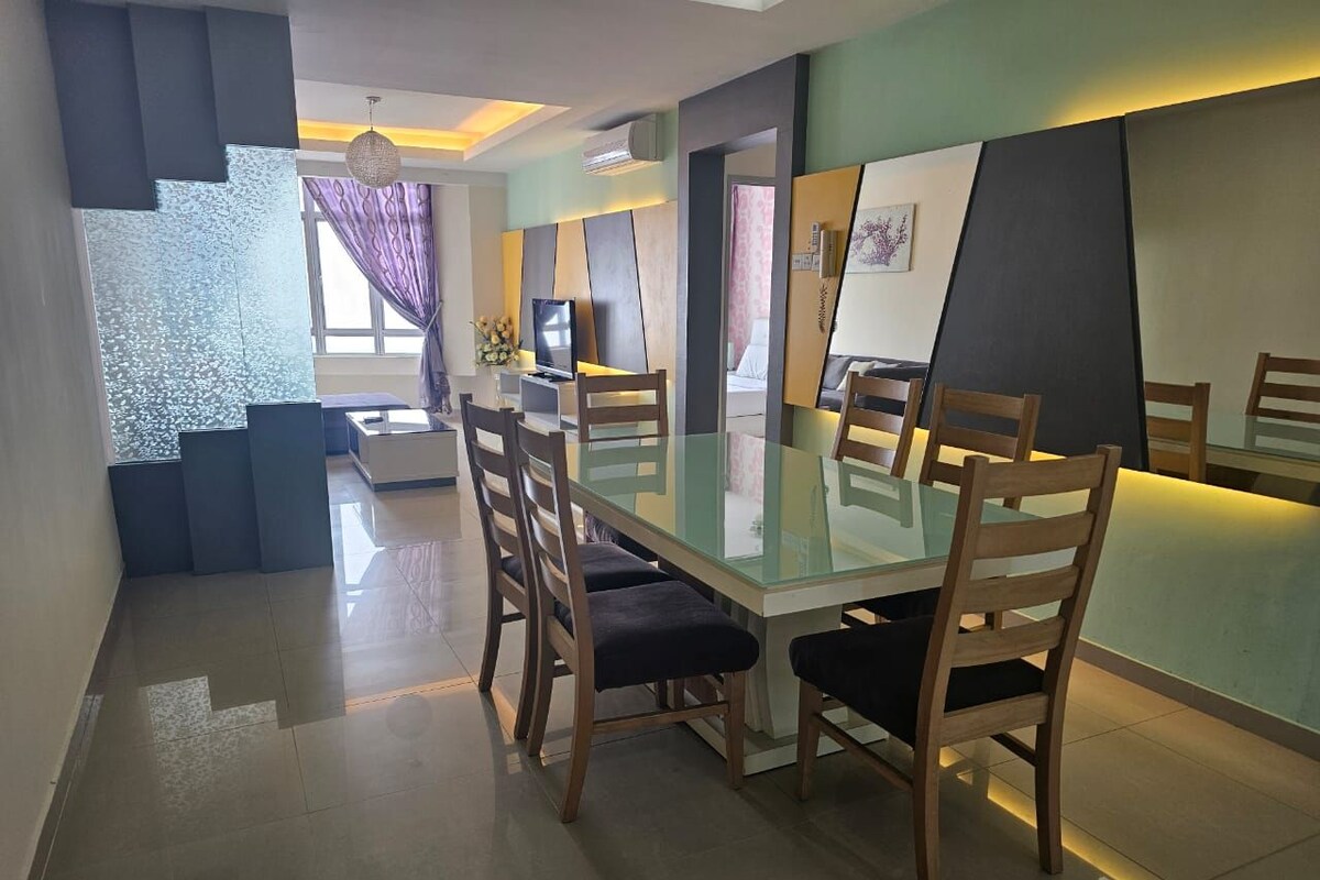 A modern dining area with a glass table and wooden chairs, a television, and a view into the living area.