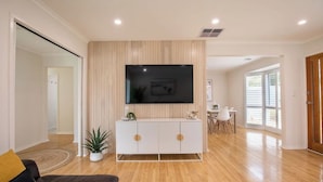 A modern living room with a flat-screen TV mounted on a wall, a white cabinet with round handles, a brown leather sofa, and a potted plant. - Ocean Breeze Retreat (Ocean Grove)
