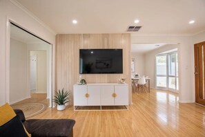 A modern living room with a flat-screen TV mounted on a wall, a white cabinet with round handles, a brown leather sofa, and a potted plant. - Ocean Breeze Retreat (Ocean Grove)