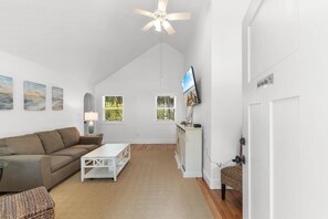 Un salon spacieux avec un plafond haut, un ventilateur de plafond, un téléviseur à écran plat fixé au mur, un canapé blanc, une table basse et un fauteuil en osier.