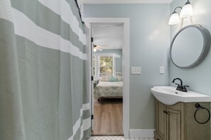 Une salle de bain avec un rideau de douche, un lavabo avec un miroir et une porte donnant sur une chambre.