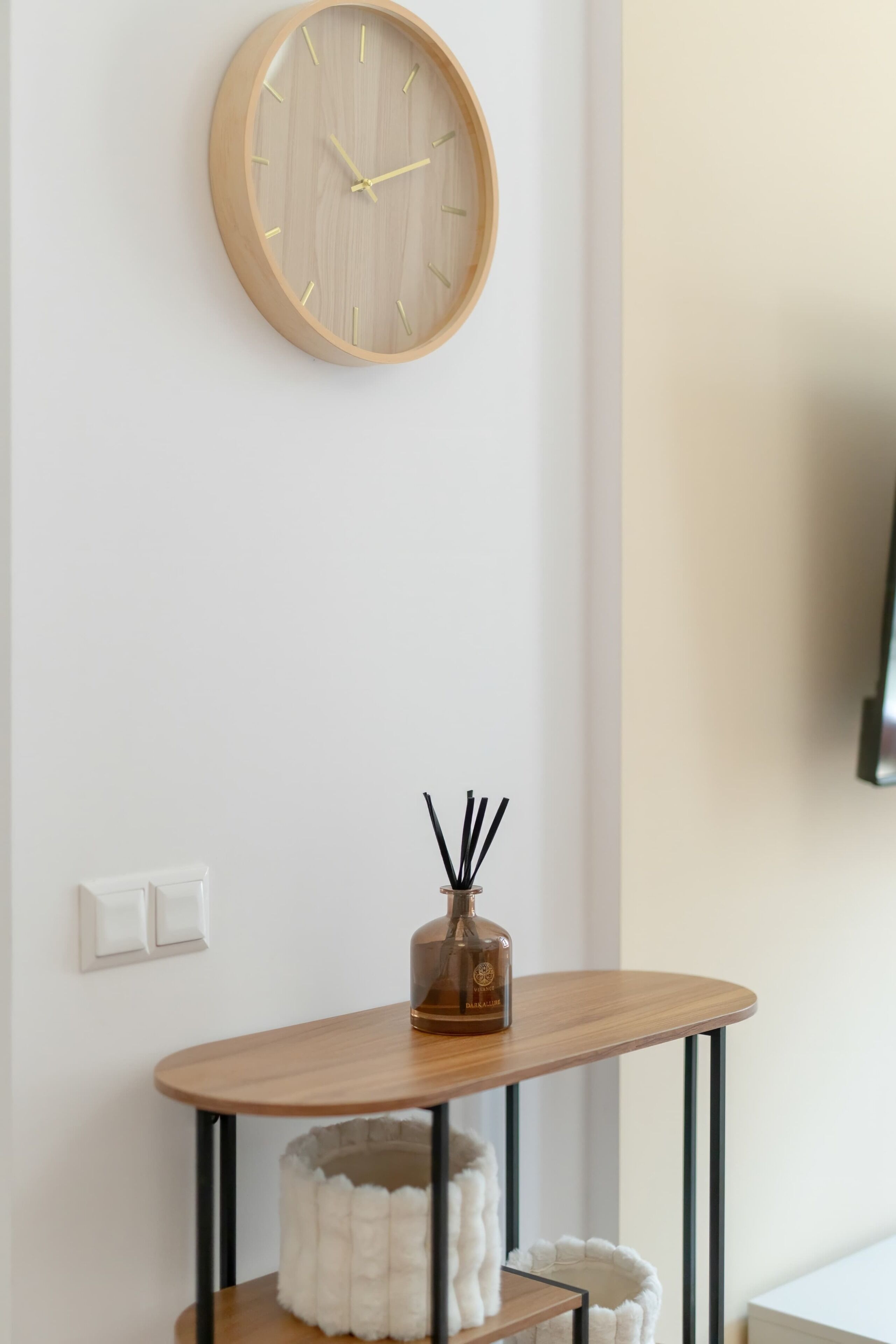 A room with a wooden wall clock, a small table with a bottle, and a white fluffy object on a shelf.