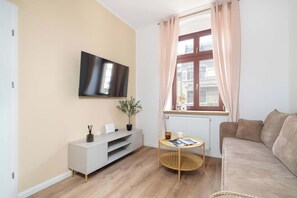 A modern living room with a flat-screen TV, a white console table, a round wooden side table, a beige sofa, and a window with curtains. - Cosy Apartments | Near the Market Square (Poznań)