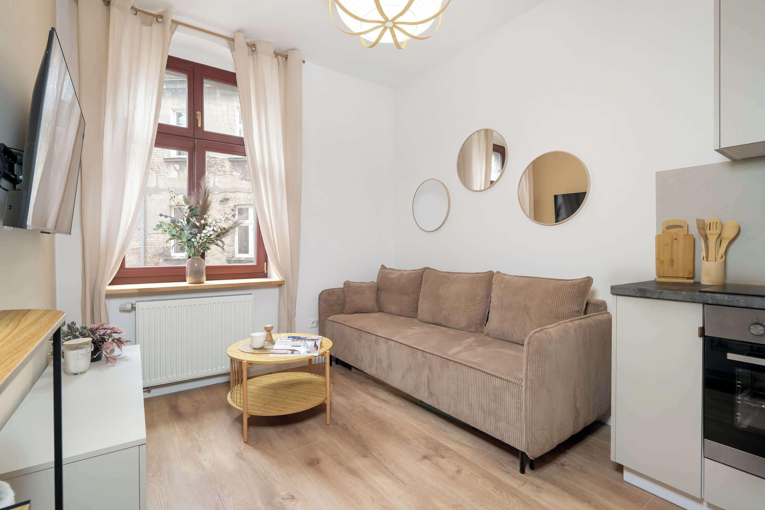 Et kompakt opholdsrum med en sofa, et lille rundt bord, et vindue med gardiner, en radiator og et tekøkken med komfur og køkkenredskaber.