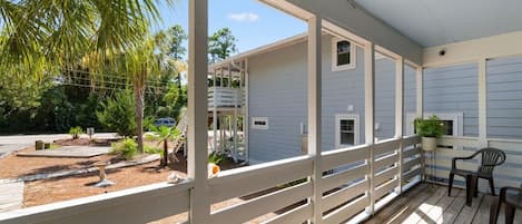 A covered porch with a white railing, a small table, and chairs, overlooking a landscaped garden with a palm tree and a building in the background.