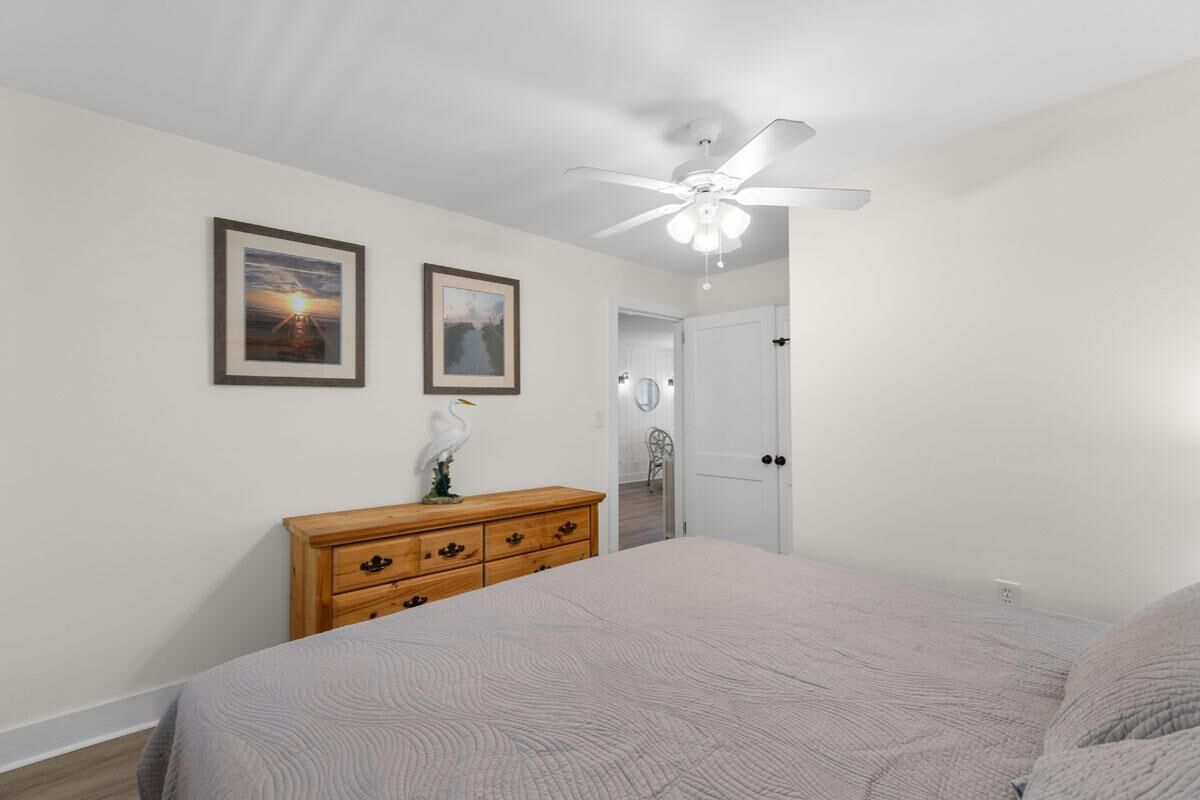 A bedroom with a bed, a dresser, two framed pictures, a ceiling fan, and a door leading to another room.