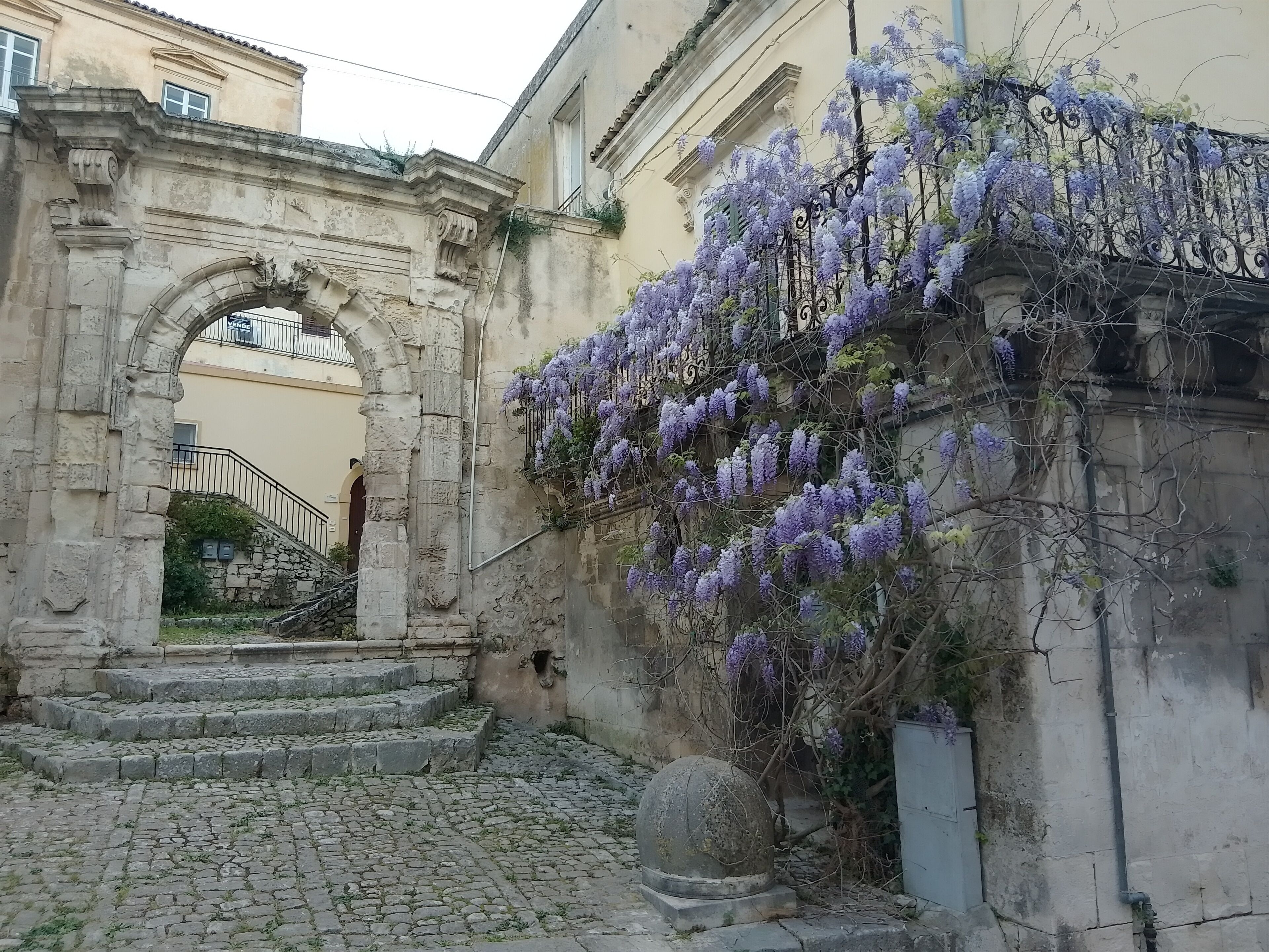 Un vecchio arco in pietra che conduce a una scala, con tralci di glicine viola che ricoprono un muro.