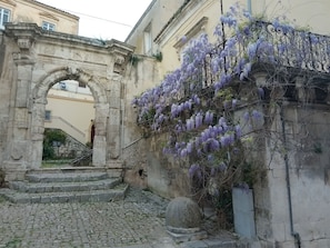 Ein alter, steinerner Bogen, der zu einer Treppe führt, mit violetten Wisteria-Ranken, die eine Mauer überwuchern.