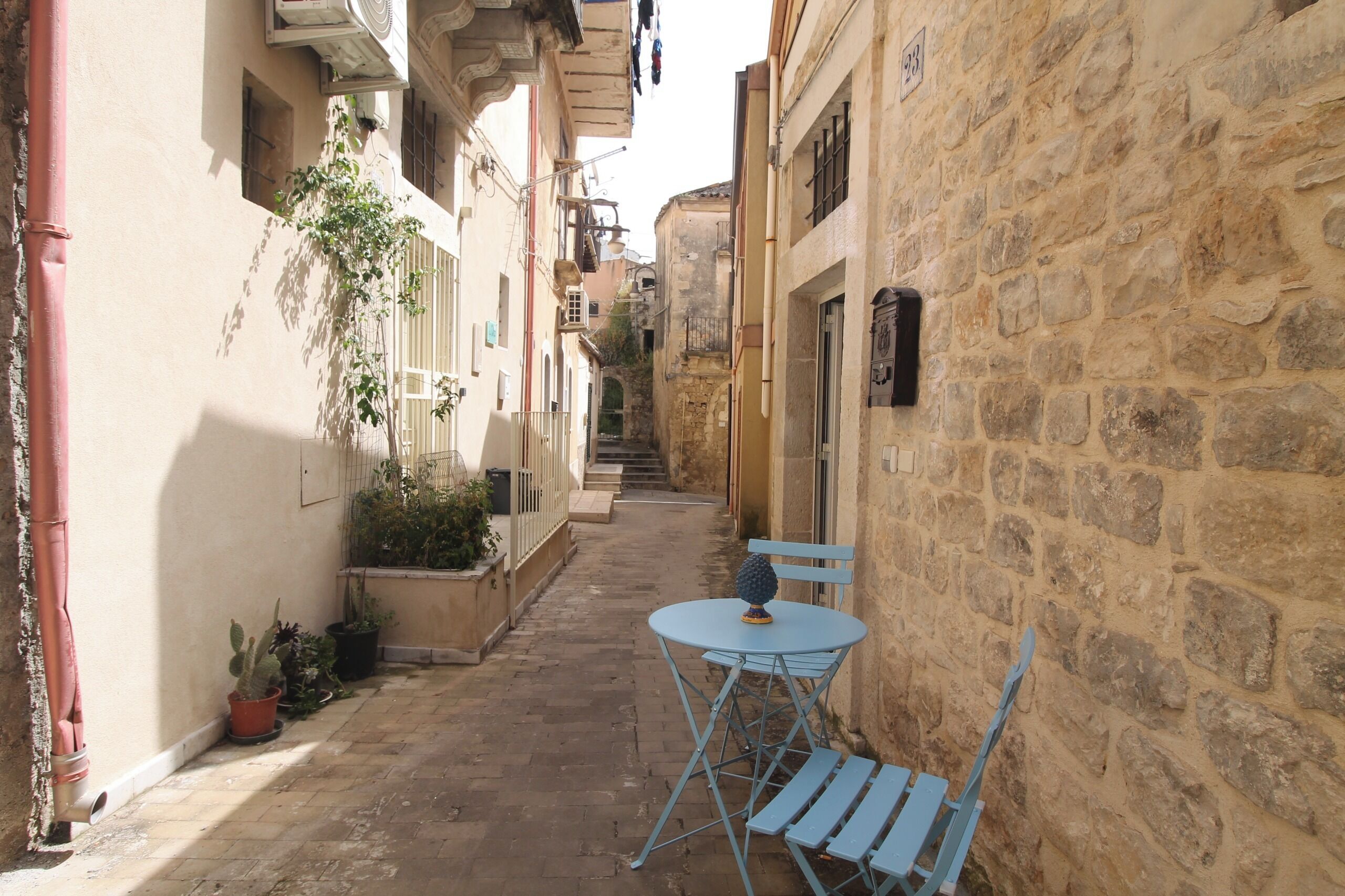 Un vicolo stretto con edifici in pietra, un tavolino con una sedia e piante in vaso.