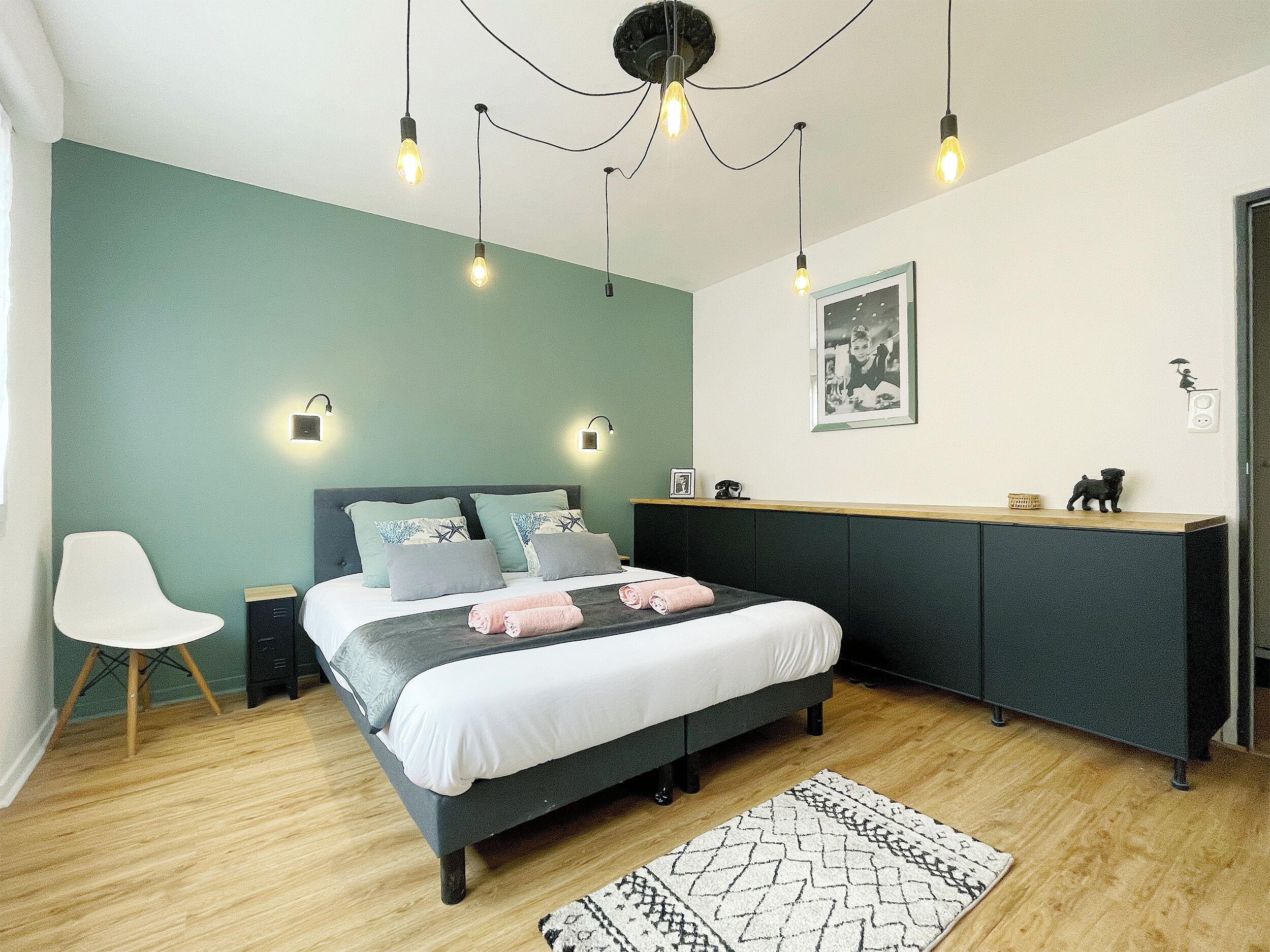 A modern bedroom with a bed, bedside tables, a chair, a desk, and a hanging light fixture.