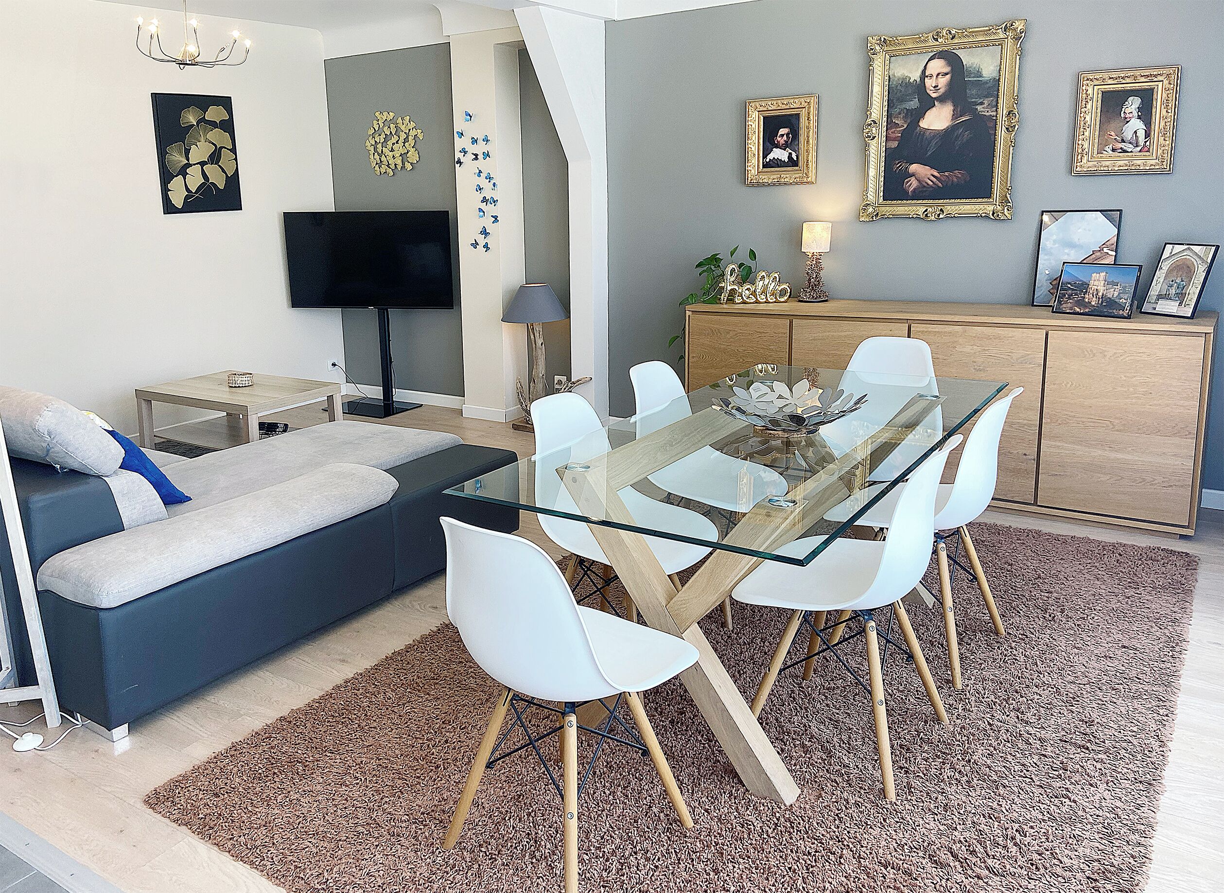 A modern living room with a glass dining table, white chairs, a sofa, a flat-screen TV, and a cabinet with framed pictures.