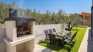 An outdoor patio with a built-in grill, a dining table with chairs, and a well-maintained lawn. - Villa Peliceti With Grand Pool (Marcana)