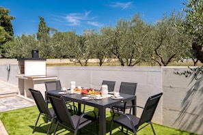 An outdoor dining area with a table set for six, surrounded by chairs, and a barbecue grill in the background. - Villa Peliceti With Grand Pool (Marcana)