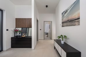 A modern hallway with a dark wooden panel, a white console table with a plant, and a painting of a sunset over the ocean. - Villa Peliceti With Grand Pool (Marcana)