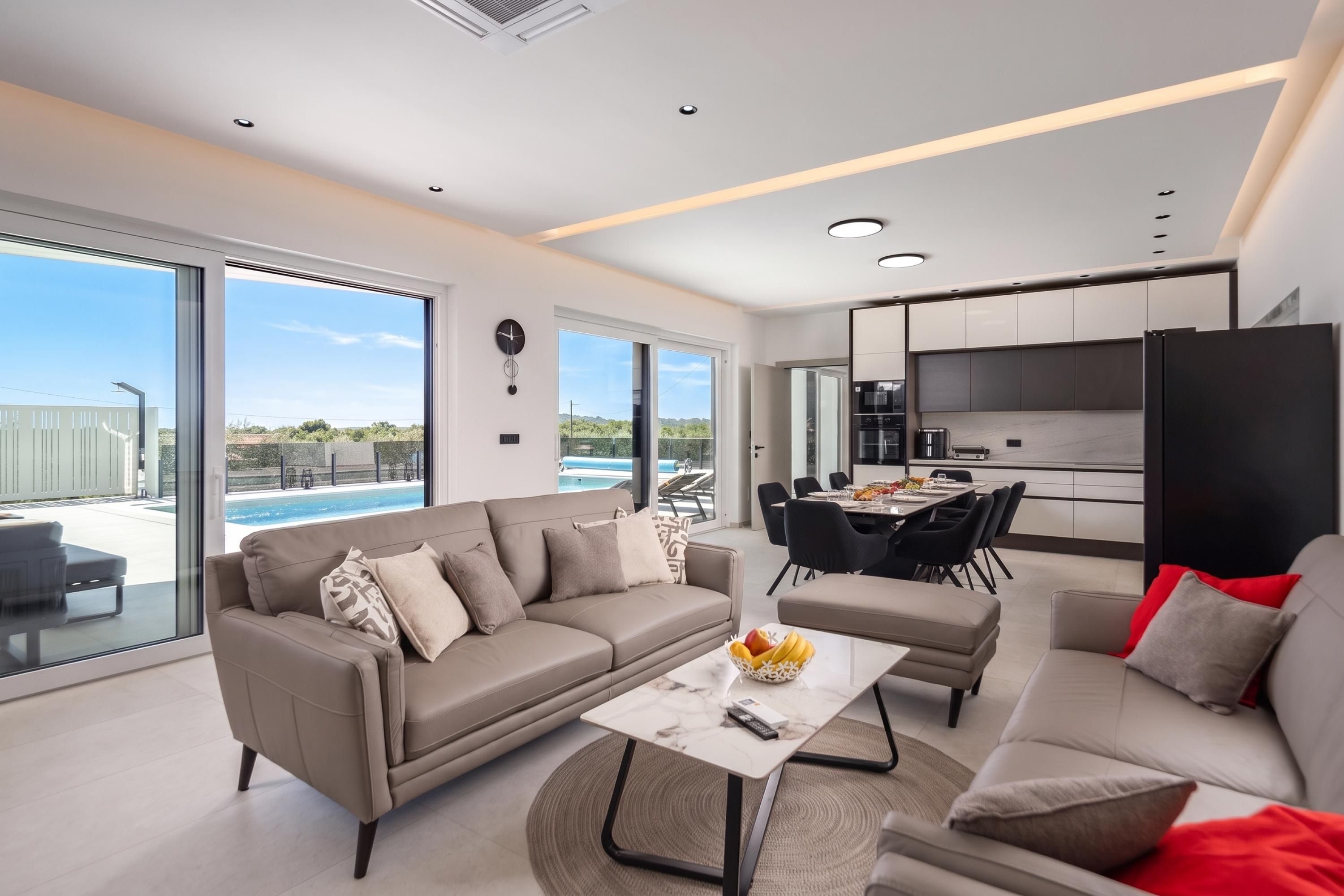 Un salon moderne avec un canapé, une table basse et un coin repas avec des chaises. Une grande fenêtre offre une vue sur une piscine.