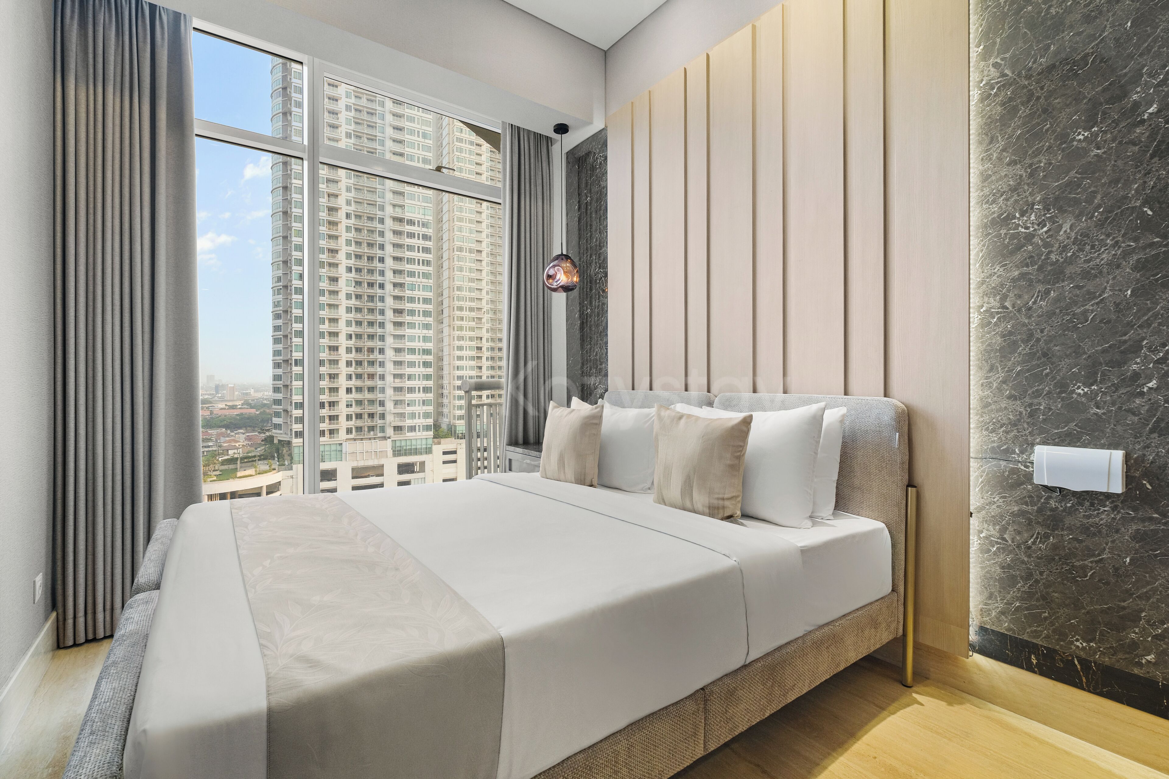 A modern bedroom with a large bed, a view of high-rise buildings, and a marble wall.