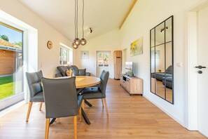 A spacious living area with a round wooden table, grey upholstered chairs, and a large window providing a view of the outdoors. - Accessible vacation home "Huus Bodderbloom" in Kating for up to 4 people (Kating)