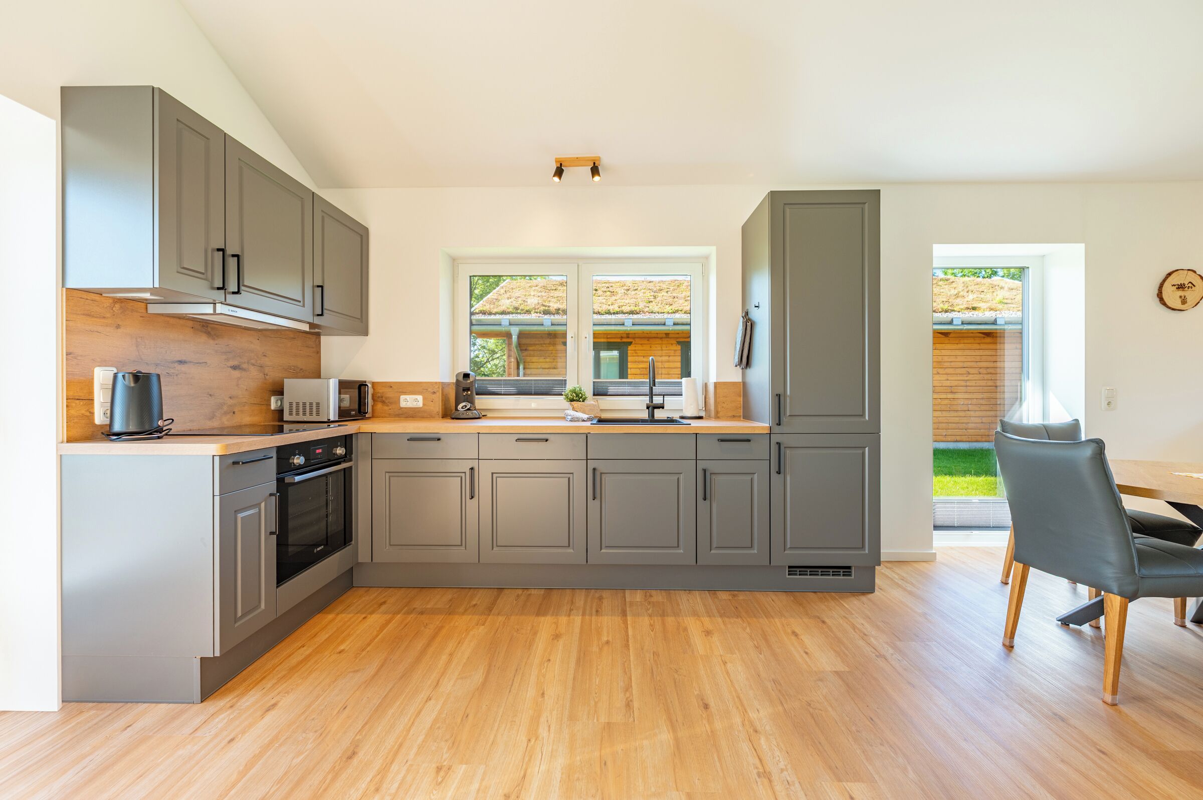 A modern kitchen with grey cabinets, a built-in oven, and a dining area with a table and chairs.