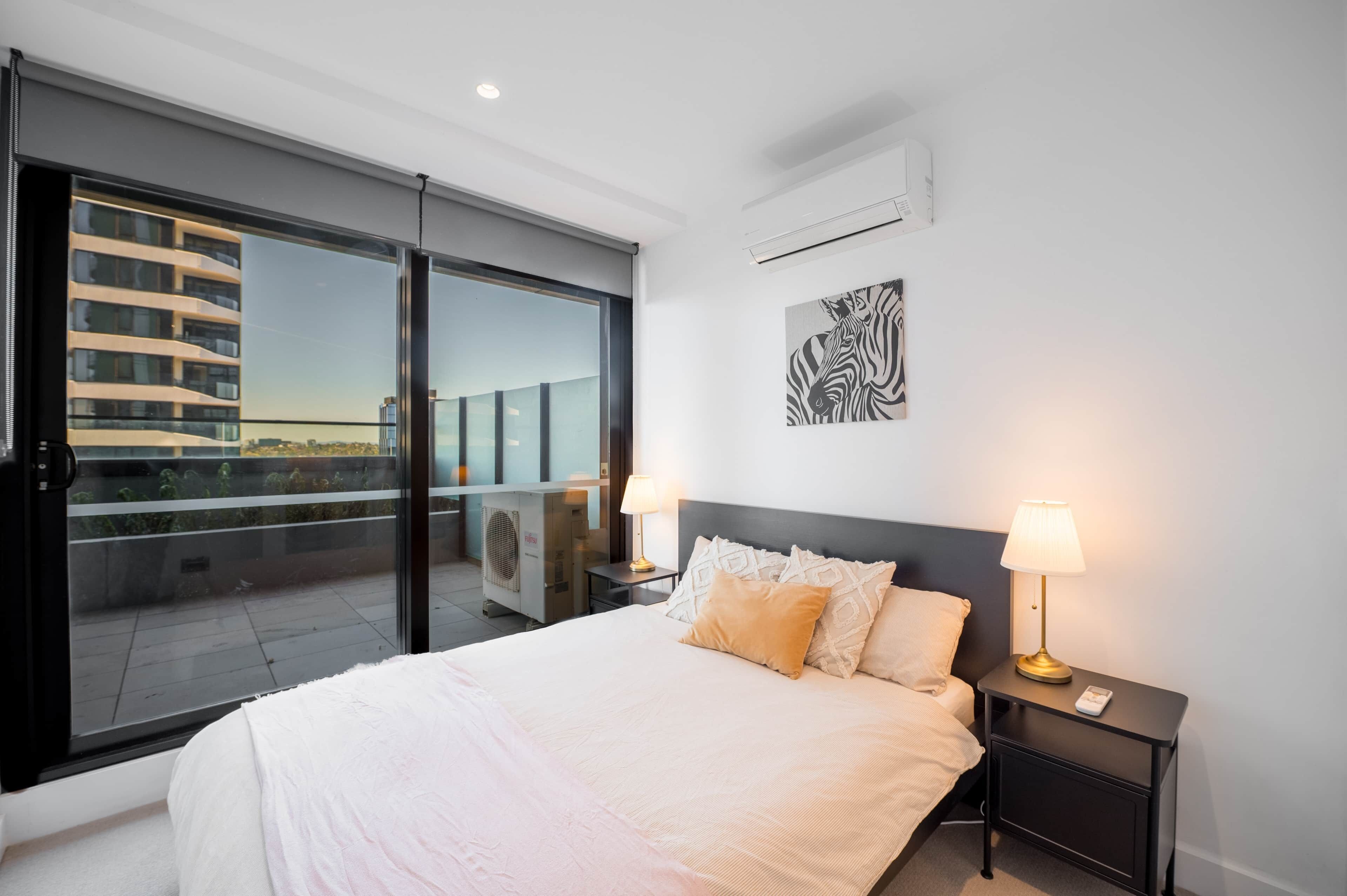 Ein Hotelzimmer mit einem großen Bett, zwei Nachttischlampen, einem Fenster mit Blick auf Gebäude und einem Deckenventilator.