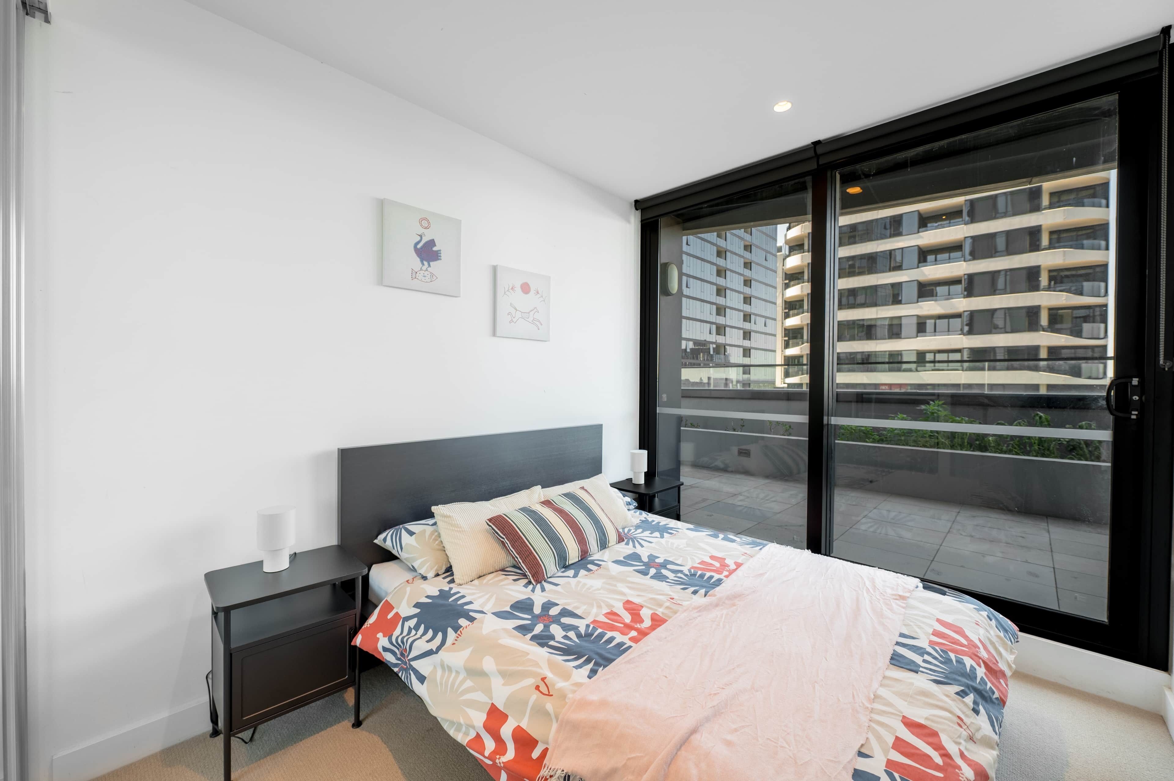 A modern bedroom with a large bed, a nightstand, a window with a view of a building, and two wall art pieces.