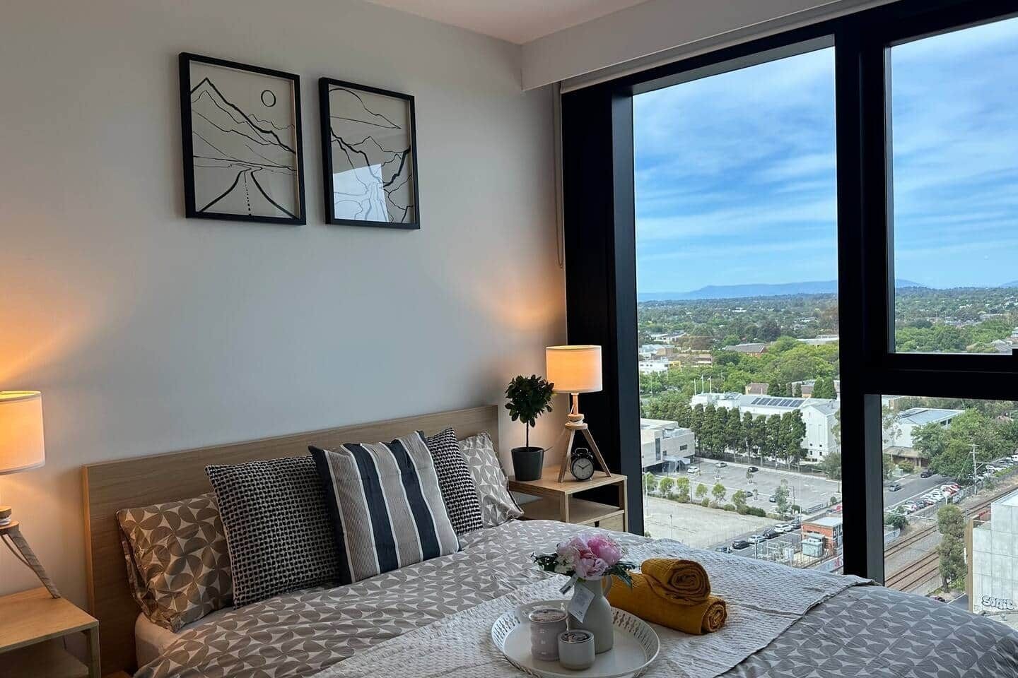 A hotel room with a large window, a bed with patterned bedding, a bedside table with a lamp, and two wall-mounted artworks.