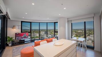 A modern living room with a large window, a flat-screen TV, and a dining area with a view of the city.