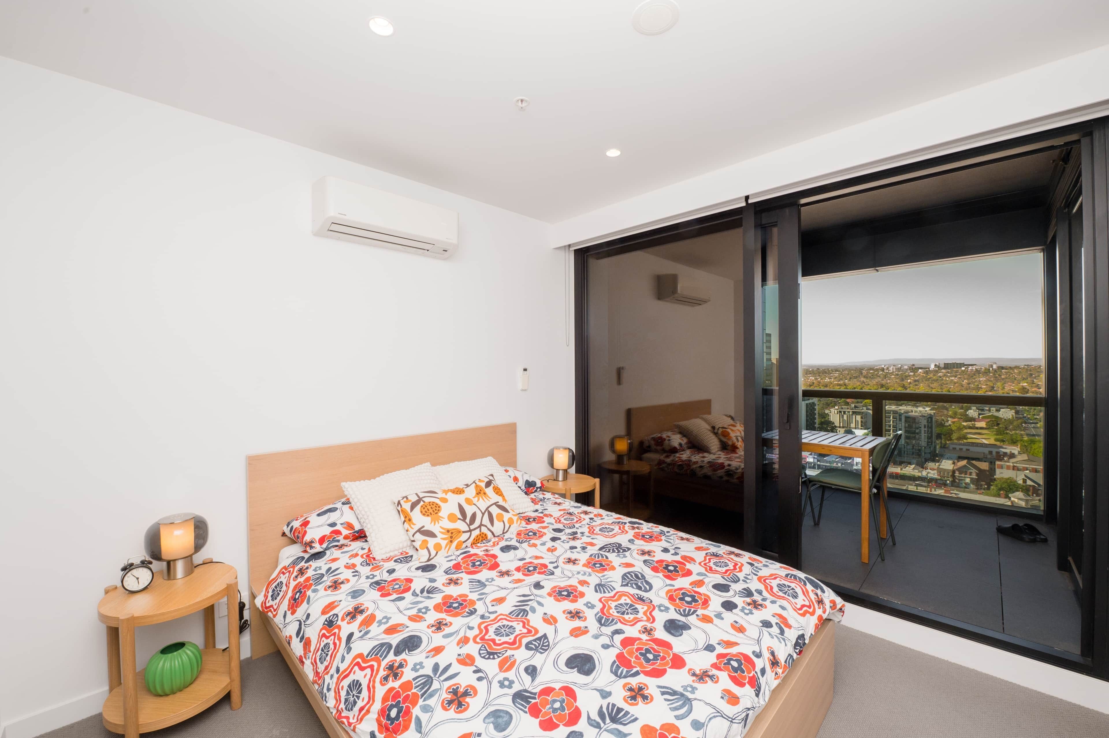 A bedroom with a bed, bedside tables, a nightstand, an air conditioning unit, and a large window with a city view.