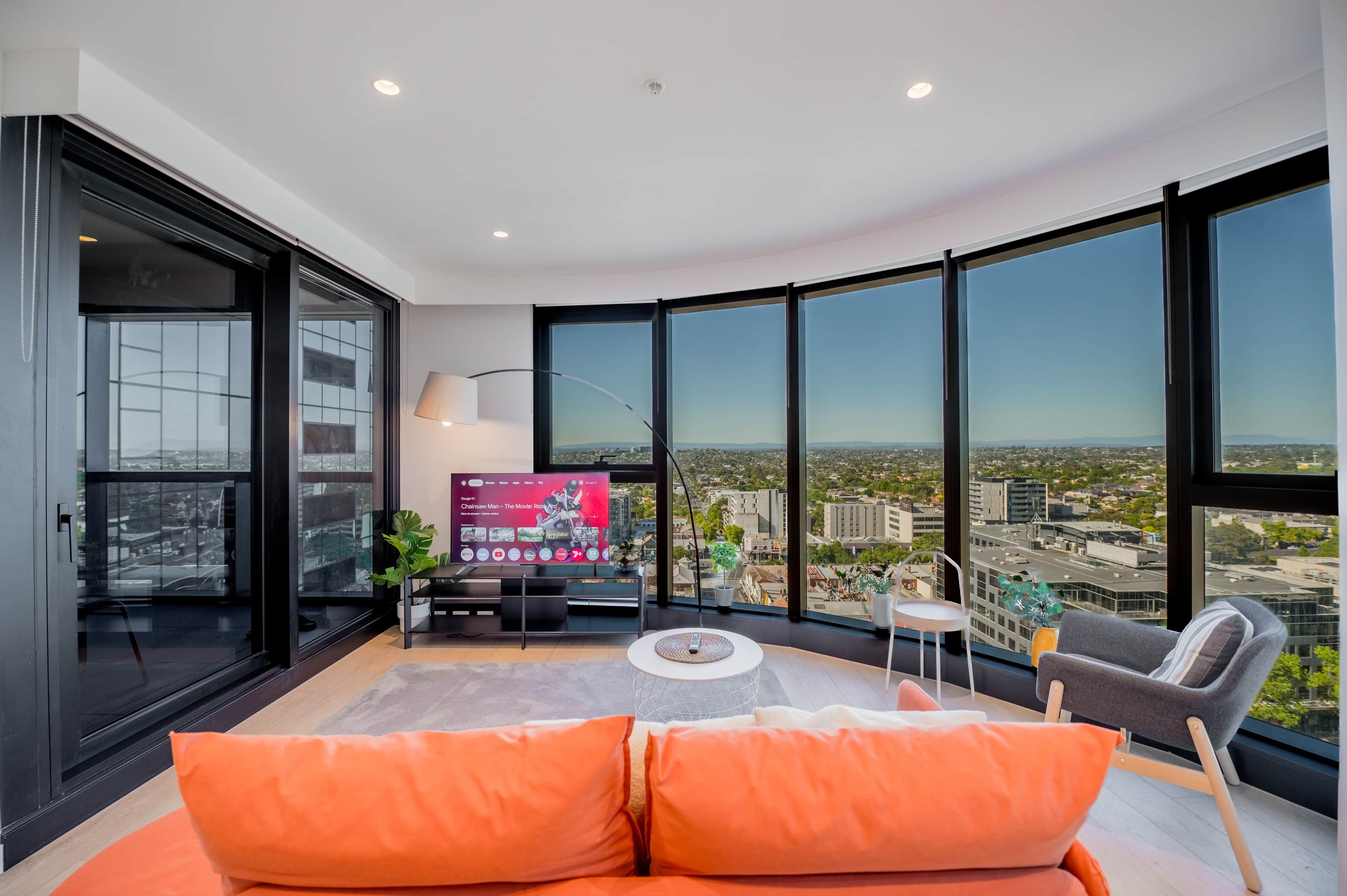 A modern living room with a large TV, comfortable seating, and a city view through large windows.