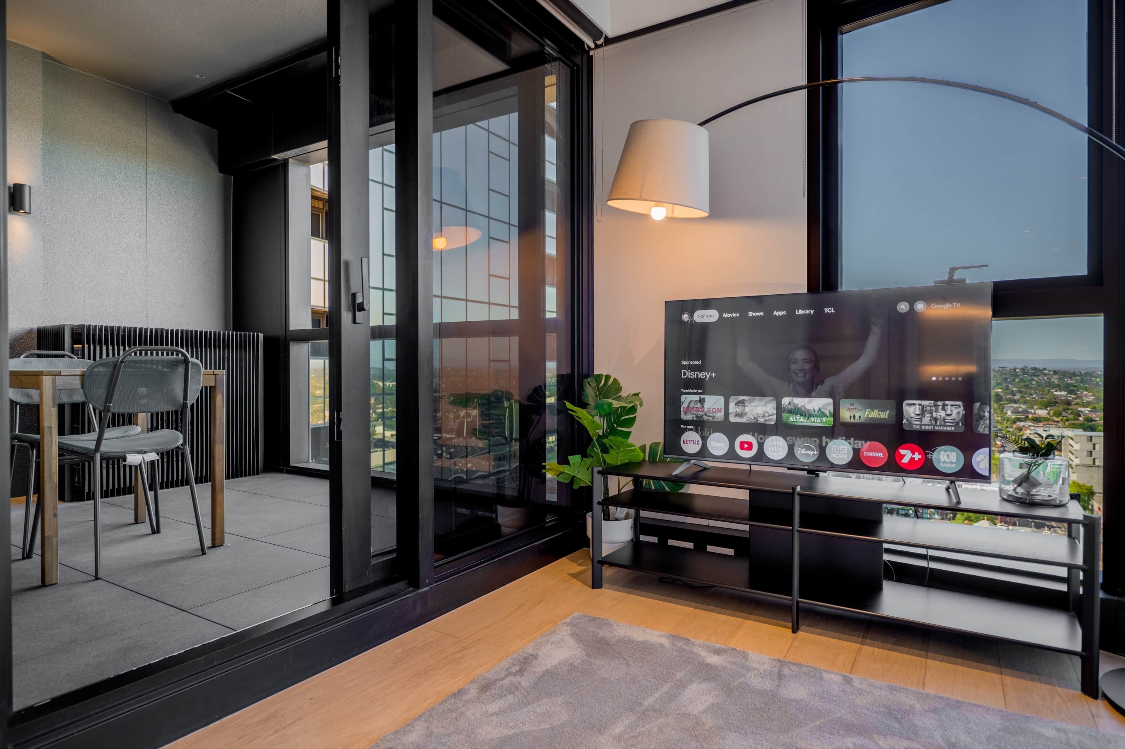 A modern living room with a large flat-screen TV, a dining area with chairs, and a view of the city through large windows.