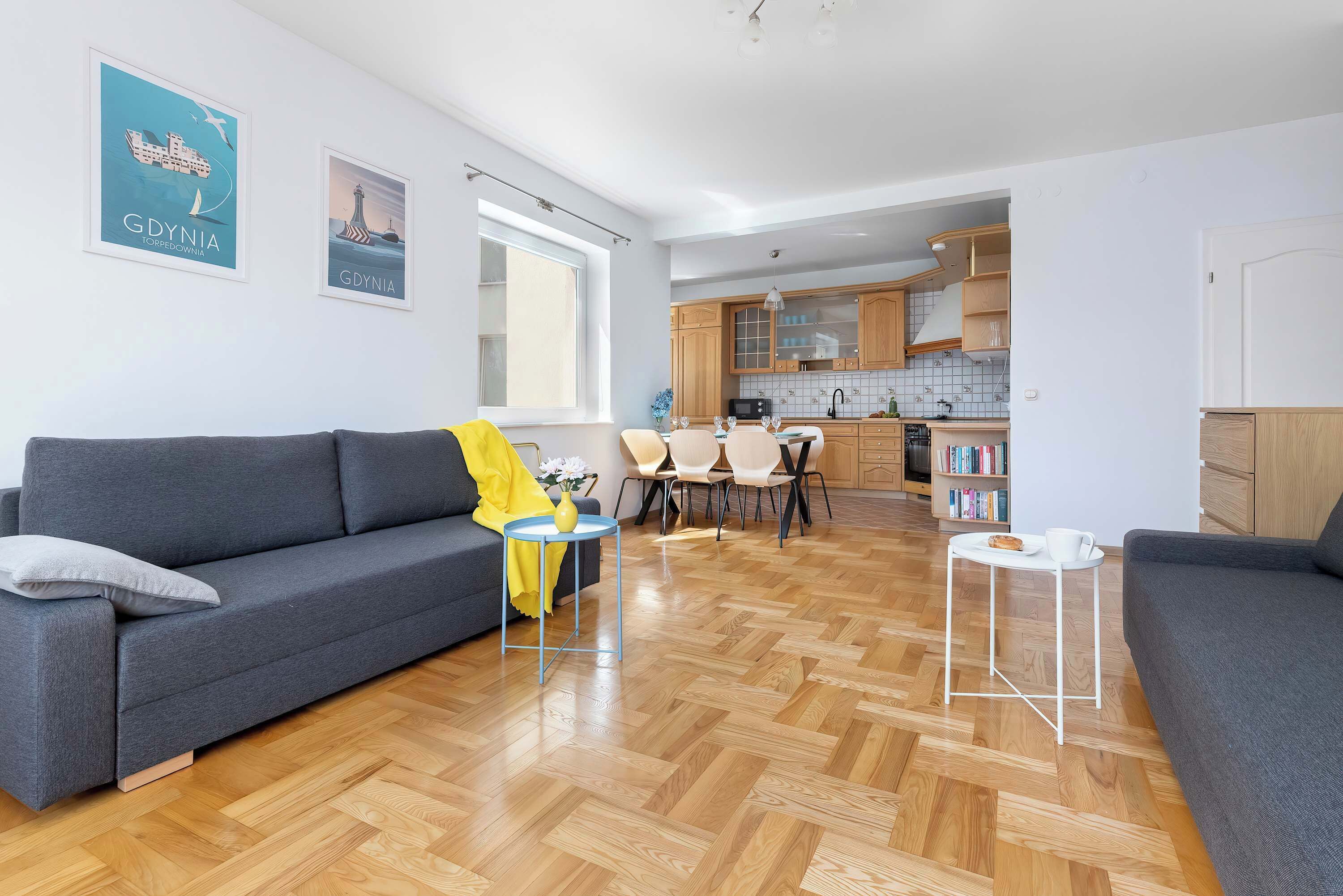 Una sala moderna con un sofá gris, una mesita redonda con un florero, y un comedor con sillas y una mesa.
