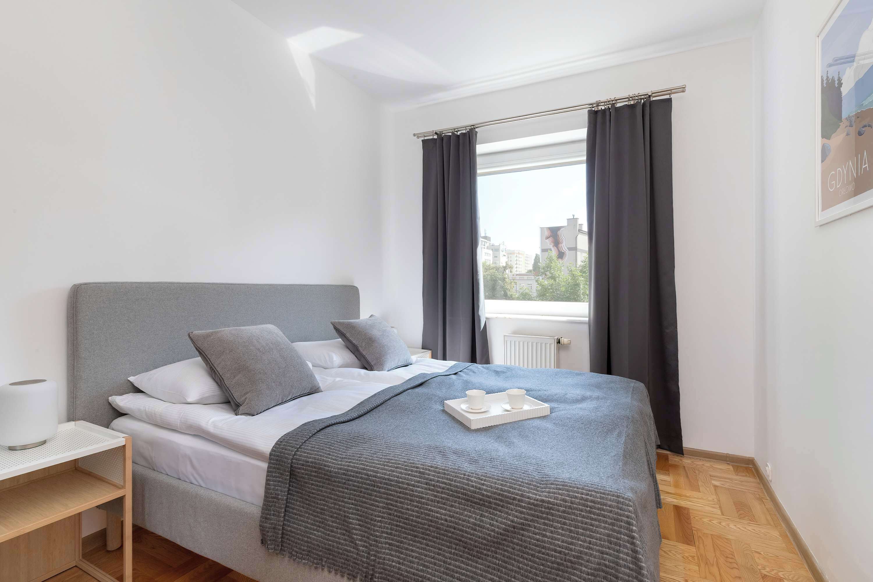 A neatly made bed with a grey headboard, white linens, and grey pillows. A grey throw blanket is draped over the bed, and a tray with two white cups is placed on the bed's footboard.