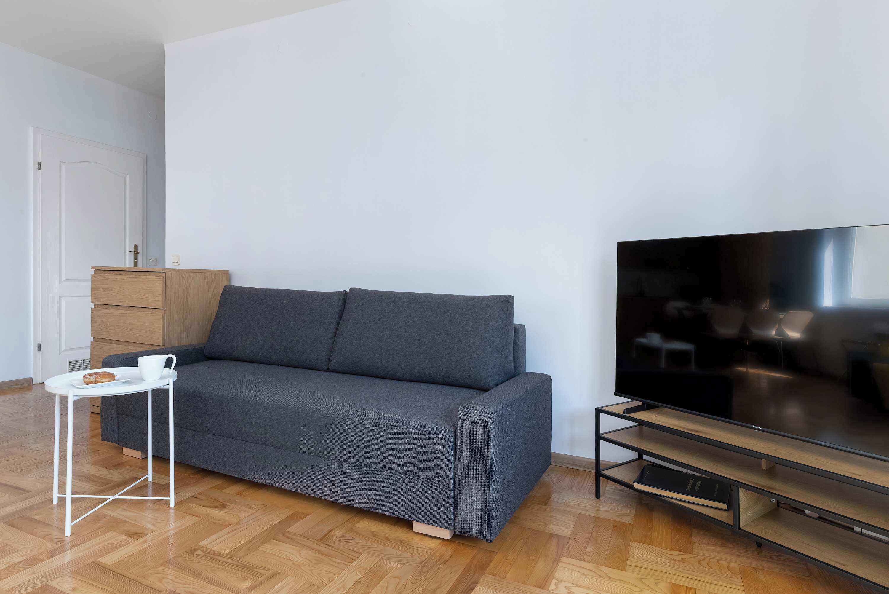 A modern living room with a grey sofa, a small white side table, a flat-screen TV, and a wooden cabinet.