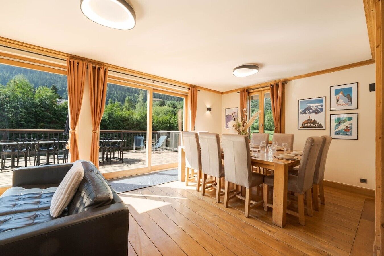 A dining area with a wooden table and chairs, large windows with curtains, and a view of greenery outside.
