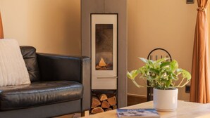 Ein Wohnzimmer mit schwarzem Ledersofa, einem weißen Zierkissen, einem Holztisch mit Fernbedienung und Buch, einer Topfpflanze und einem Kamin mit Kaminfeuer.