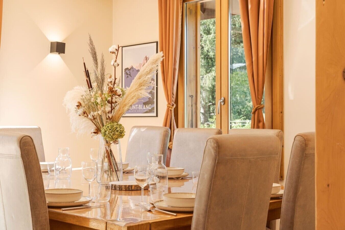 A dining area with a table set for four, chairs, a vase with dried flowers, a framed picture on the wall, and a window with curtains.