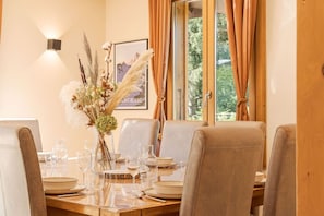 Un coin repas avec une table dressée pour quatre personnes, des chaises, un vase avec des fleurs séchées, un tableau encadré au mur et une fenêtre avec des rideaux.