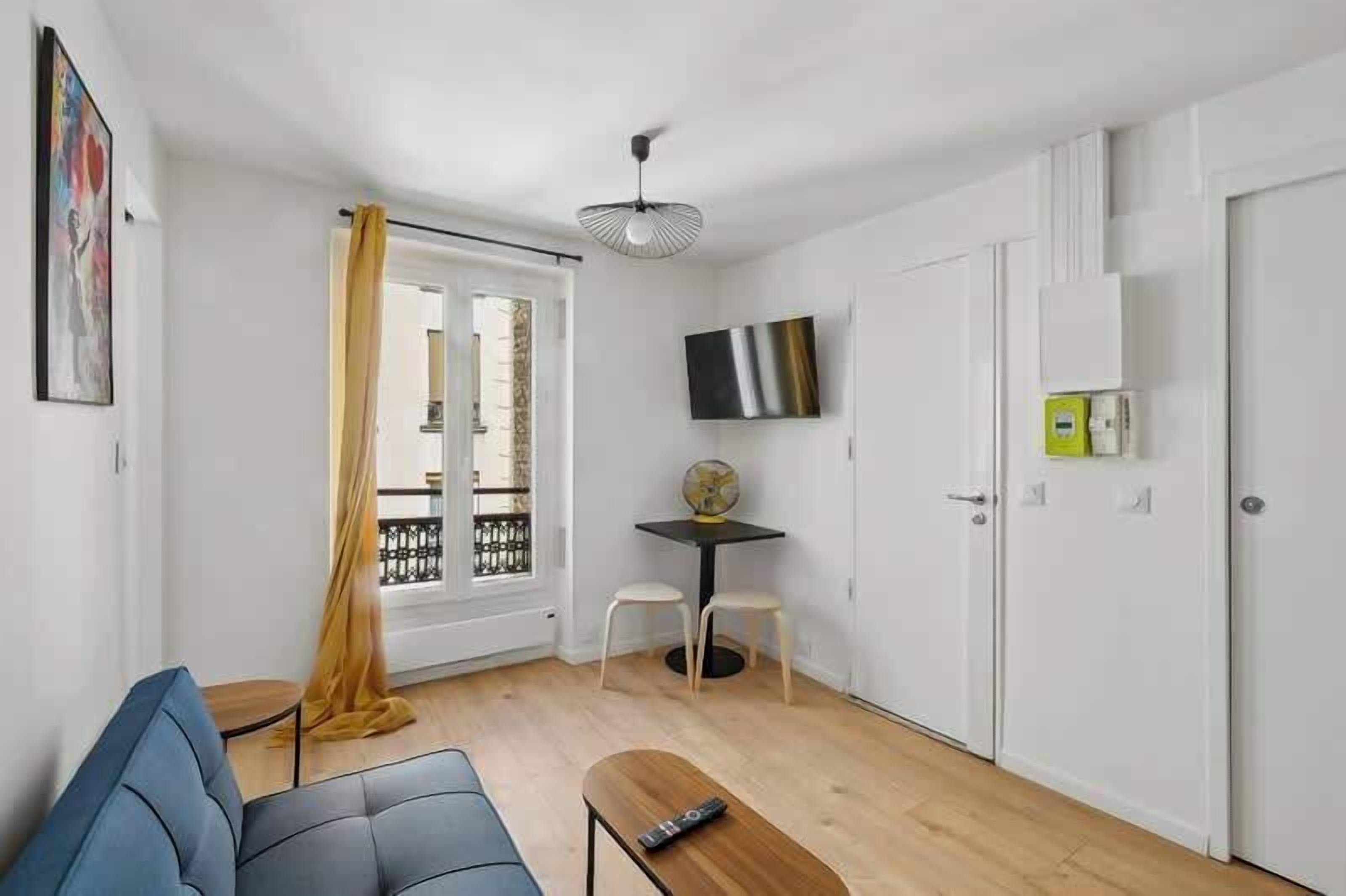 A compact living space with a sofa, a small table, a TV, and a balcony with a yellow curtain.