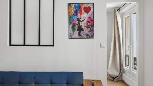 Een kamer met een blauwe bank, een klein tafeltje, een kleurrijk kunstwerk en een raam met gordijnen.