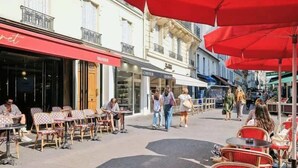 Een openluchterras met tafels en stoelen, een rode luifel, en mensen die over het trottoir lopen.