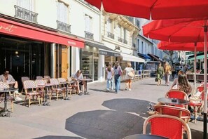 Ein Straßencafé mit Tischen und Stühlen, einer roten Markise und Passanten auf dem Bürgersteig.