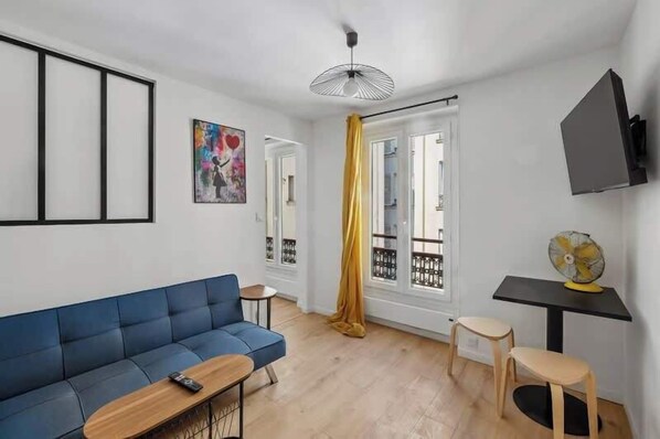 A living room with a blue sofa, a small table, a fan, and a TV mounted on the wall. - Central and Bright 1BR- Near Parc Monceau (Paris)