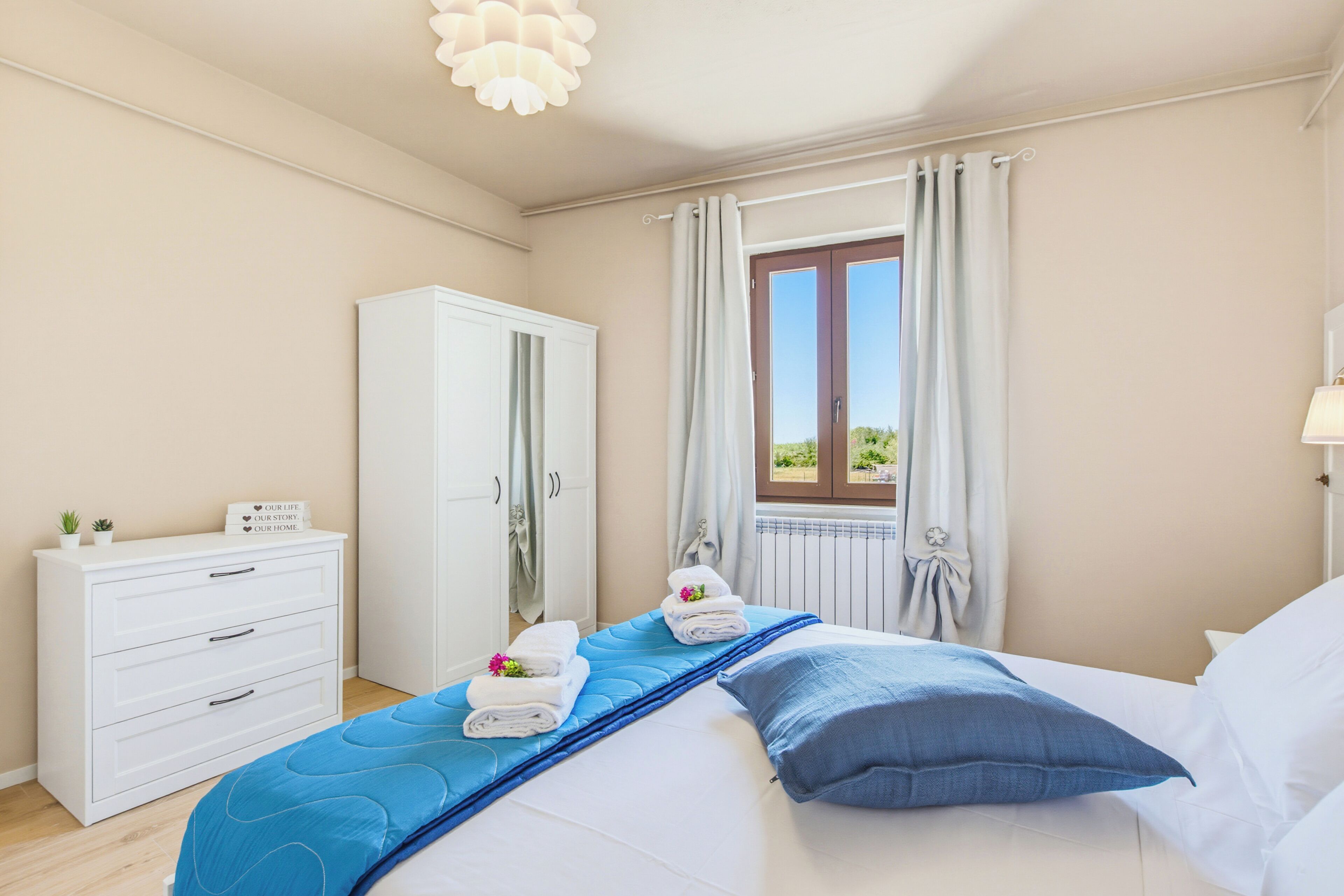 Ein Schlafzimmer mit einem Bett, einem Kleiderschrank, einem Fenster mit Vorhängen und Blick ins Grüne.