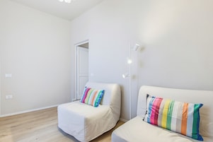 Habitación minimalista con dos sofás blancos, cada uno con un cojín de rayas coloridas, una lámpara de techo blanca y suelo de madera.