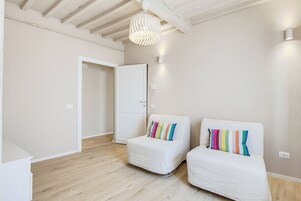 Una stanza con pavimento in legno, pareti bianche, una porta bianca e due divani bianchi decorati con cuscini colorati.