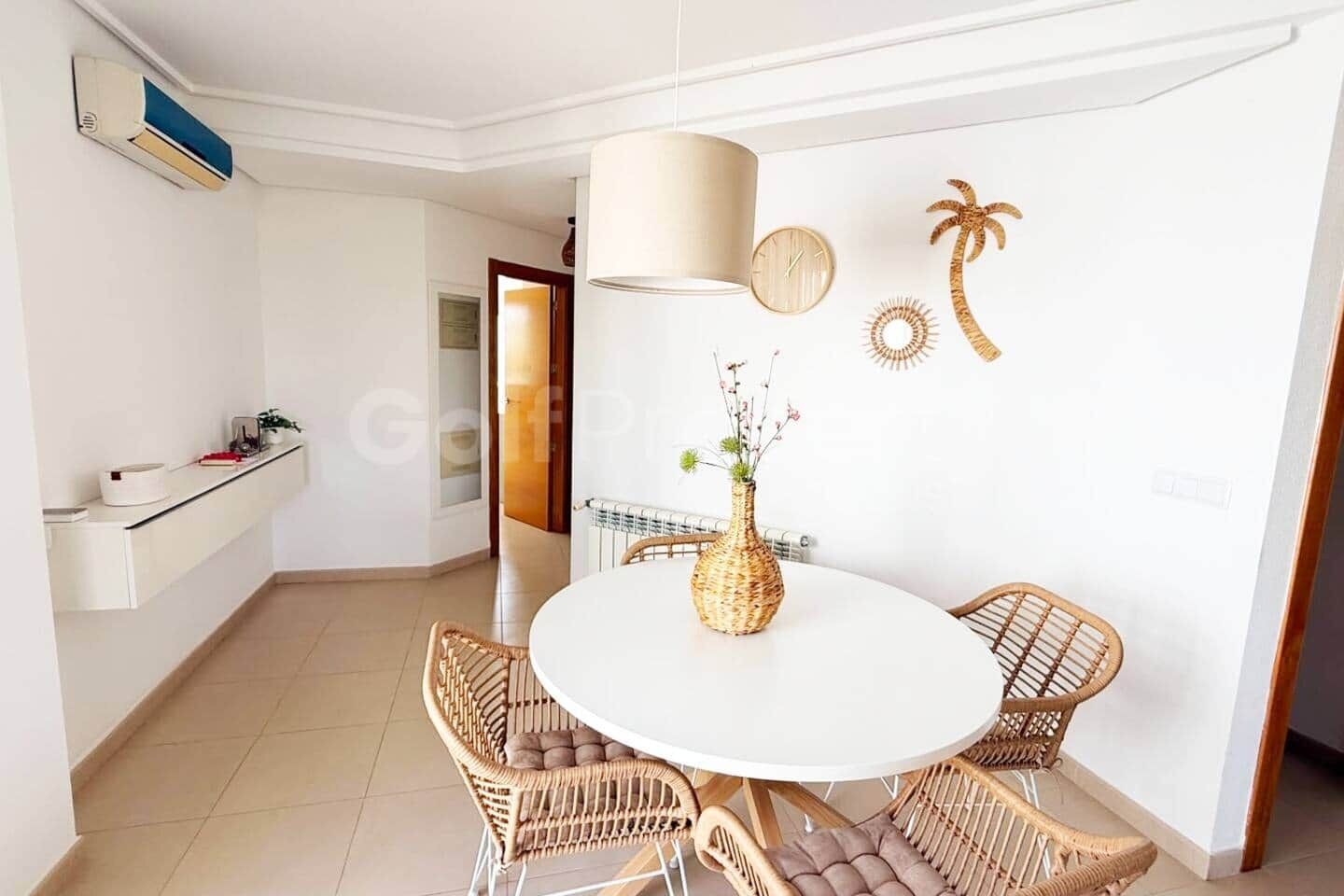 Un comedor con una mesa redonda, sillas de mimbre, un florero con flores, un reloj de pared y un aire acondicionado de pared.
