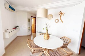 A dining area with a round table, wicker chairs, a vase with flowers, a wall clock, and a wall-mounted air conditioner. - Well Furnished West-Facing 2 Bed 1 Bath Apartment (Sucina)